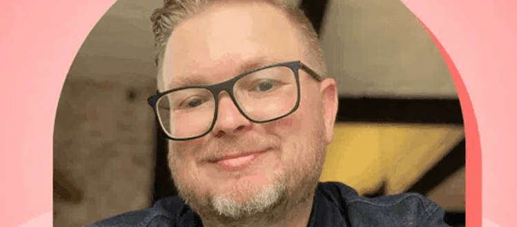 A person wearing black-framed glasses and a dark shirt smiles warmly at the camera in a close-up selfie photo